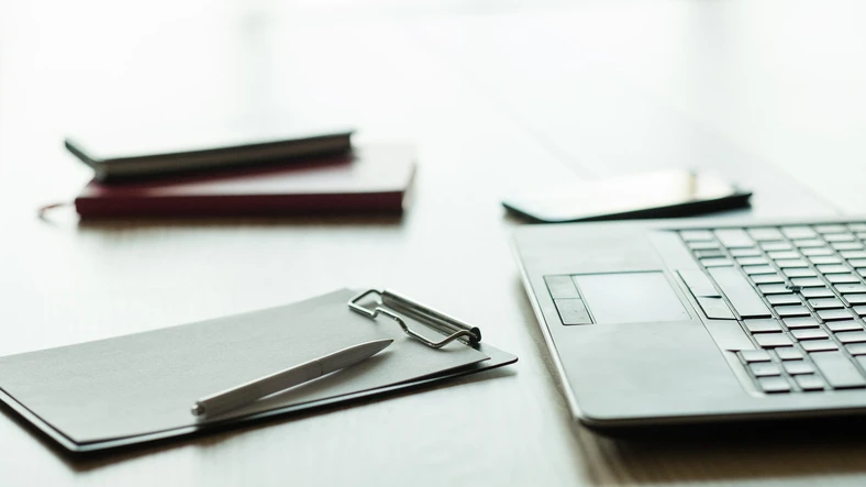 Very bright picture of a work desk with soft reflections and a blurry, almost vanishing, background.
                  Within the scene there's a business laptop, a smartphone, a pen, two notebooks and a clipboard.
